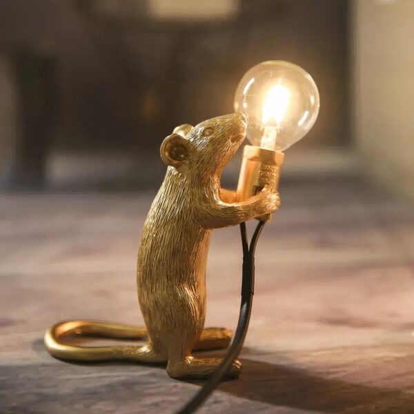 Lampe de chevet en forme de souris dorée sur ses deux pattes arrière tenant une ampoule allumée entre ses pattes avant. Sur une surface couleur bois. L'ampoule est relié à un fil.