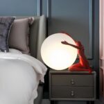 Lampe de chevet en forme de lune allumée avec un humanoïde rouge assis à droite et tenant la lune. La lampe est posée sur une table de chevet grise à 2 tiroirs. A sa gauche, un lit avec tête de lit grise et drap blanc. Le mur derrière est bleu.