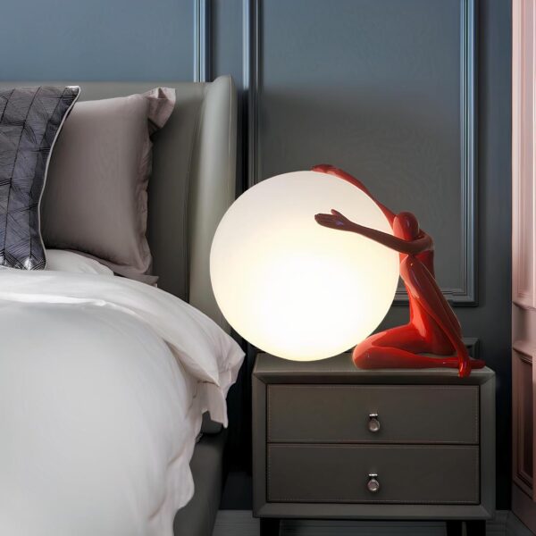 Lampe de chevet en forme de lune allumée avec un humanoïde rouge assis à droite et tenant la lune. La lampe est posée sur une table de chevet grise à 2 tiroirs. A sa gauche, un lit avec tête de lit grise et drap blanc. Le mur derrière est bleu.