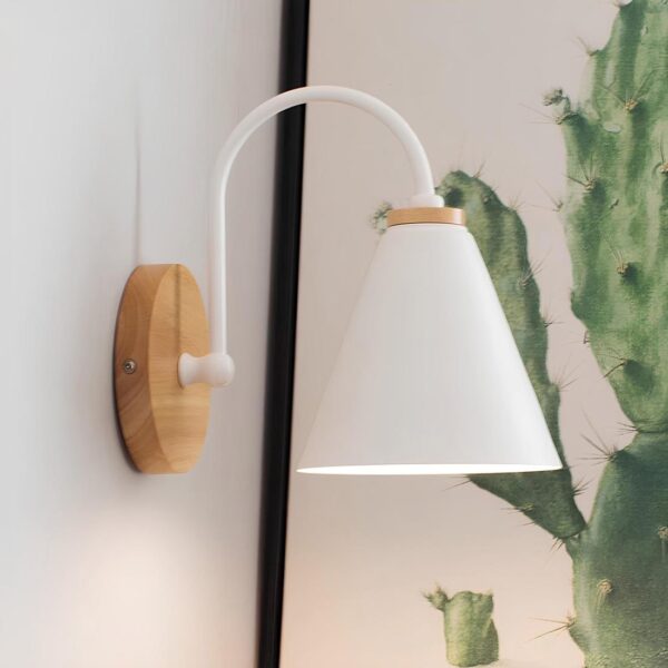 Lampe de chevet murale de style scandinave avec support rond en bois au mur et cône blanc en métal tenu par une tige arrondie reliée au support en bois. Lumière allumée, un cactus en arrière-plan à droite.