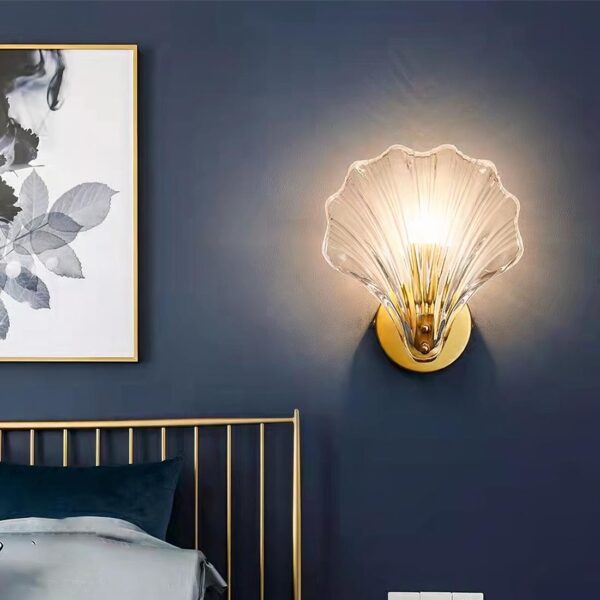Lampe de chevet murale en forme de coquillage transparent en verre sur socle rond doré fixé au mur. L'ampoule est allumée. La lampe est fixée sur un mur bleu foncé. A gauche un lit en métal doré avec literie bleue. Au-dessus, un cadre blanc et gris.