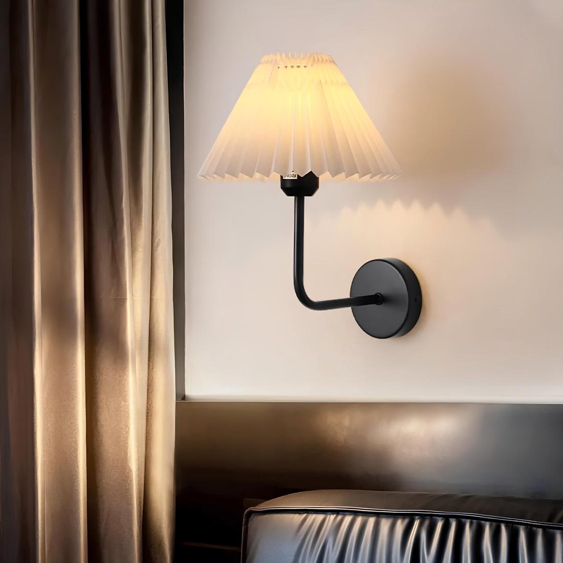 Lampe de chevet murale avec socle rond noir fixé au mur et tige coudée vers le haut noire. Par-dessus, un abat-jour blanc avec une ampoule allumée. A gauche un rideau marron. En dessous une partie du mur marron et un coussin de fauteuil en simili cuir marron.