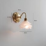 Lampe de chevet murale vintage avec socle rond motifs de pétales de fleurs dorées avec tige en col de cygne dorée. Un abat-jour en verre représentant une fleur. L'ampoule est allumée. Fixée sur un mur gris.