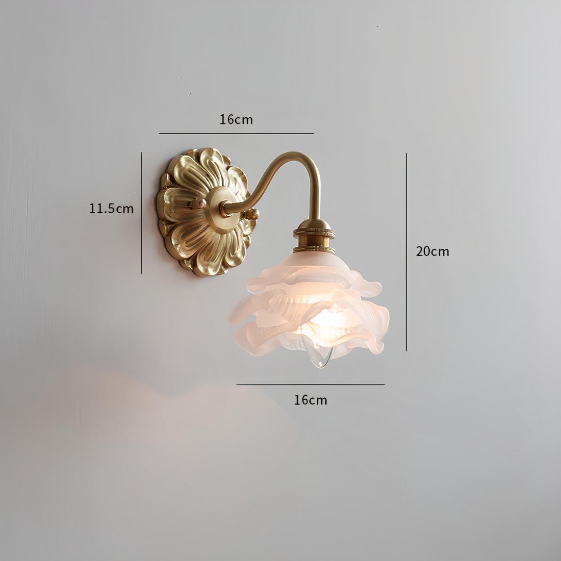 Lampe de chevet murale vintage avec socle rond motifs de pétales de fleurs dorées avec tige en col de cygne dorée. Un abat-jour en verre représentant une fleur. L'ampoule est allumée. Fixée sur un mur gris.