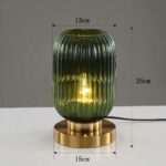 Lampe de chevet style vintage de forme cylindrique arrondi en verre strié vert avec ampoule allumée à l'intérieur. Le verre est posé sur un socle doré de forme ronde. La lampe est posée sur un support blanc, le fond est gris. Les dimensions sont affichées autour de la lampe.