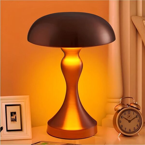 Lampe de chevet de couleur rose dorée avec une lumière blanc chaud en forme de champignon avec le corp évasé. A droite un petit réveil à aiguilles et un rideau blanc derrière. A gauche un cadre blanc.