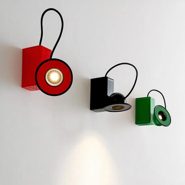 3 lampes de chevet murales alignées sur un mur blanc. De gauche à droite : rouge, noir et vert. Les lampes sont identiques. Une partie rectangulaire fixée au mur, un fil noir part du dessus jusqu'à un spot lumineux aimanté à la partie rectangulaire. Le sport est vu de face, le noir éclaire vers le bas, le vert vers le haut.