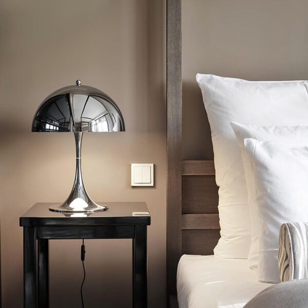 Lampe de chevet en forme de champignon de couleur argentée posée sur une table de chevet noire. A droite, un lit avec des oreillers blancs. Le mur derrière est couleur taupe. Lampe est allumée et filaire avec un interrupteur.