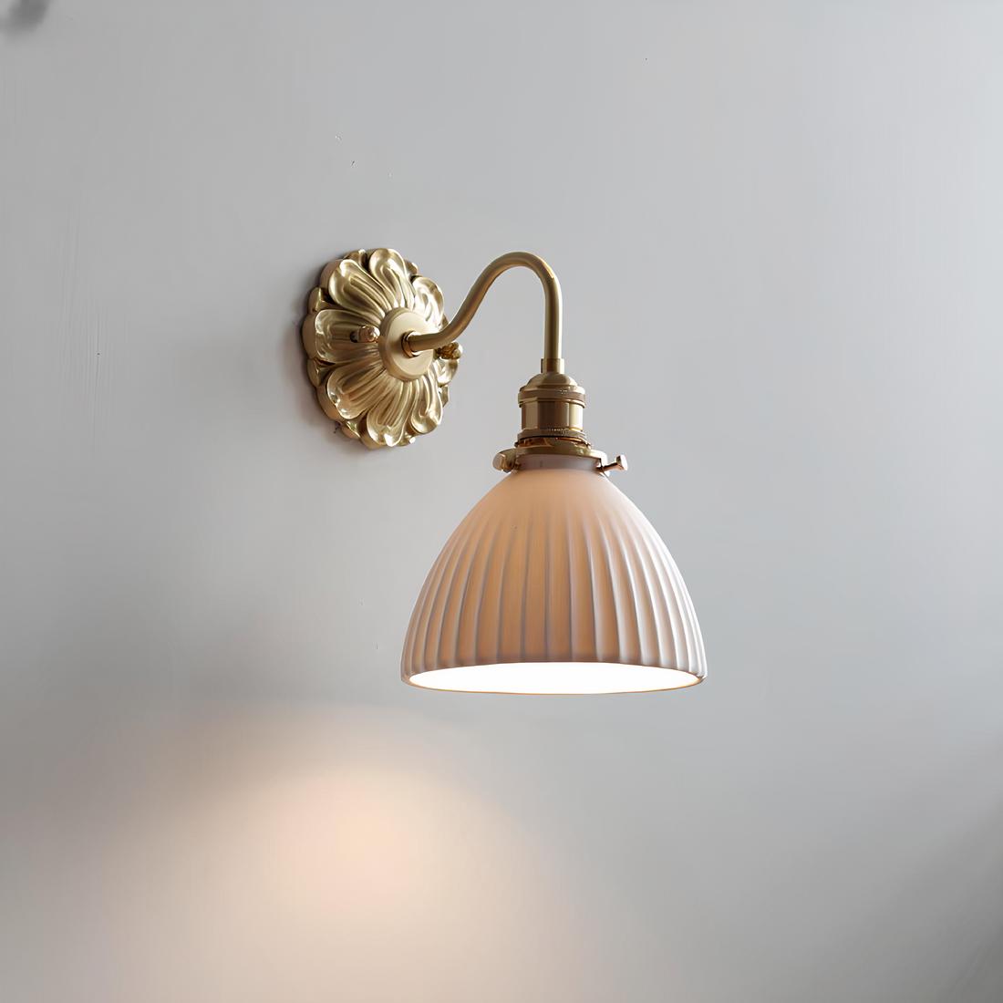 Lampe de chevet murale avec socle rond doré travaillé avec des motifs imitant des pétales de fleurs, une tige en col de cygne vers le bas dorée avec un abat-jour en forme de cloche en céramique blanche. L'ampoule est allumée.