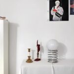 Lampe de chevet en forme de ressort sur lequel est posée une boule lumineuse allumée reliée à son câble. Posée sur un support couleur blanc. A sa droite une affiche en noir et blanc