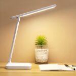 Lampe de lecture blanche sans fil pliante avec tête lumineuse longue. Posée sur un support couleur bois clair au-dessus d'un cahier avec stylo, une petite plante artificiel en fond devant un mur beige.