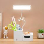 Lampe de lecture blanche avec rangements pour stylos. Flexible orientable, tête lumineuse LED longue. Ciseaux, feutres et correcteur dans le rangement de gauche, gomme à droite. A gauche un sujet en forme d'astronaute. A droite une mini plante artificielle. Mur beige. Posée sur un support couleur bois clair.