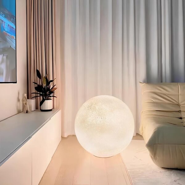 Lampe de chevet posée au sol en forme de lune allumée éclairant ses caractéristiques de la surface de la lune. A droite, un canapé beige. Derrière, un grand rideau plissé blanc. A gauche, un long meuble bas, blanc avec une plante verte. Le sol est du parquet flottant beige.