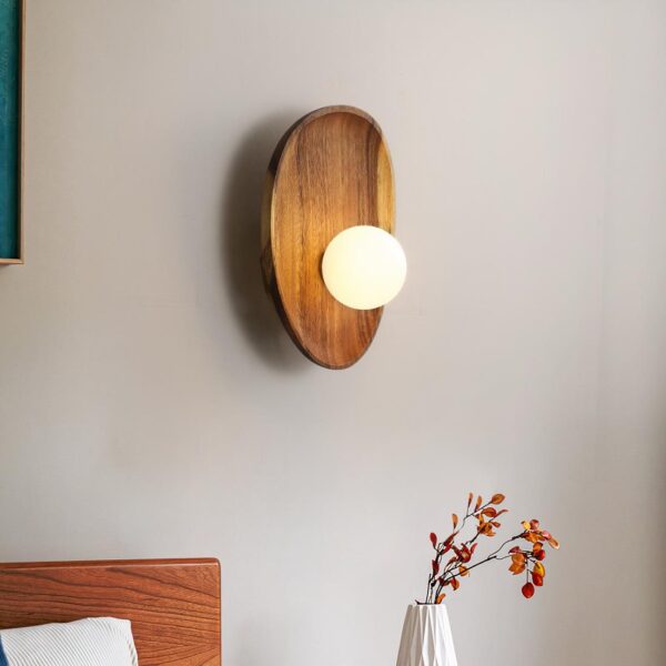 Lampe de chevet murale de forme ovale en bois naturel avec une boule lumineuse dessus. Fixée au-dessus d'un vase blanc avec des fleurs fines roses, une tête de lit à gauche.