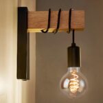 Lampe de chevet murale de style rétro industriel. Rectangle en métal noir fixé au mur sur lequel est fixé un rectangle en bois perpendiculaire. Un câble noir est enroulé sur le bois pour laisser suspendre la douille noire et l'ampoule ronde dont on voit le filament allumé.