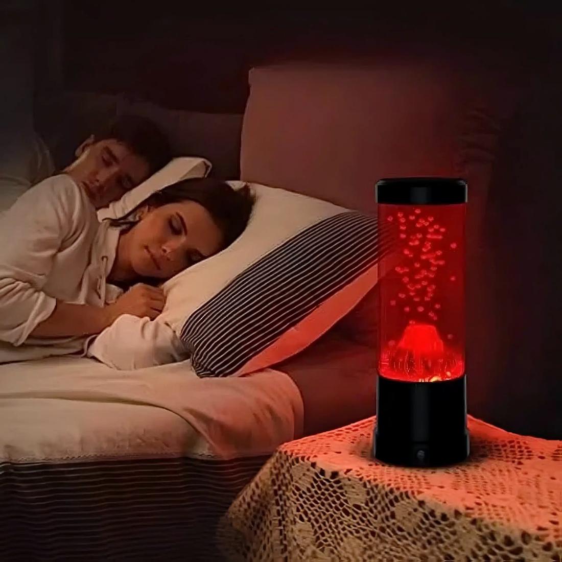 Une lampe à lave effet eruption de volcan posée sur une table de chevet avec un couple en arrière plan qui dort