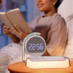 Une lampe de chevet réveil avec chargeur rapide intégré posée sur une table de chevet avec en arrière plan une personne qui lit dans son lit