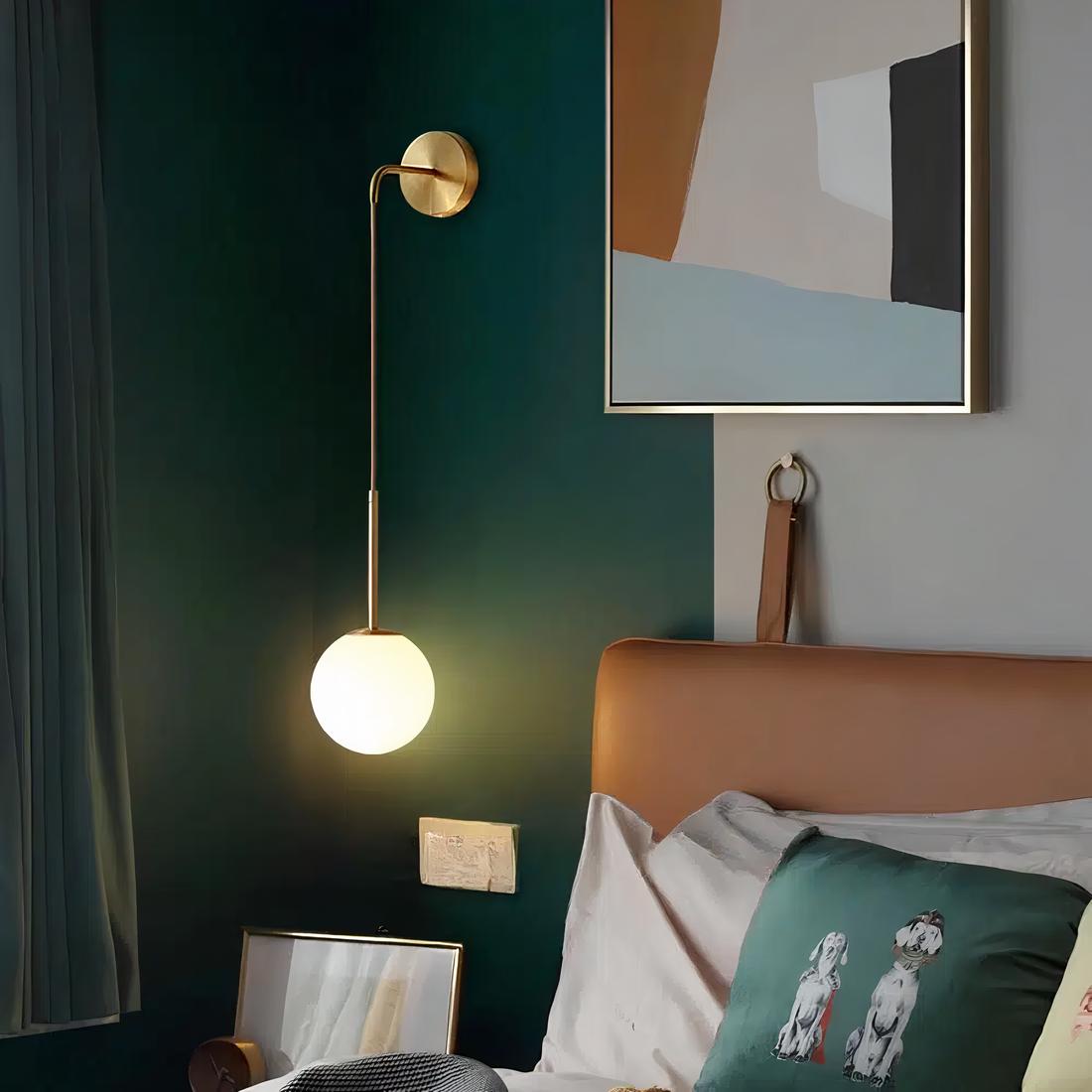 Lampe de chevet suspendue de couleur dorée fixée sur un mur vert foncé. La lampe est allumée.A droite, on retrouve un lit avec une tête de lit marron et un coussin vert sur un coussin blanc.Au dessus du lit, un cadre abstrait aux couleurs pastel.