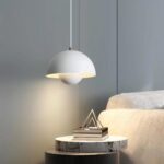 Lampe de chevet suspendue de couleur blanche avec un design nordique, la lampe est allumée. Elle est fixée au-dessus une petite table de chevet ronde, le mur derrière est gris. A droite, un canapé beige.