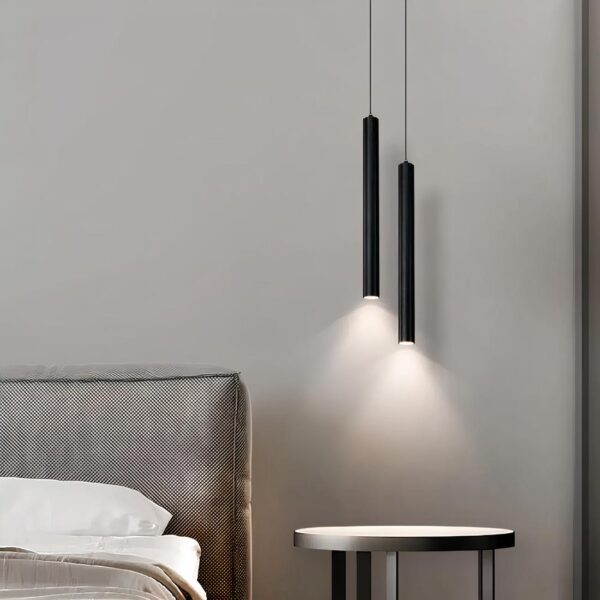 Lampe de chevet suspendue en forme de tube noir. Deux tubes noirs tenus par un câble et éclairant vers le bas. En dessous, une table ronde de chevet avec à sa gauche un lit gris et blanc.