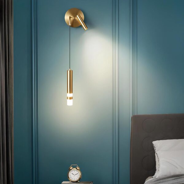 Lampe murale suspendue de couleur dorée avec un tube dont le bout est lumineux. Sur le même socle mural, une liseuse dorée dirigée vers le lit qui est à droite. Fixée sur un mur bleu canard.