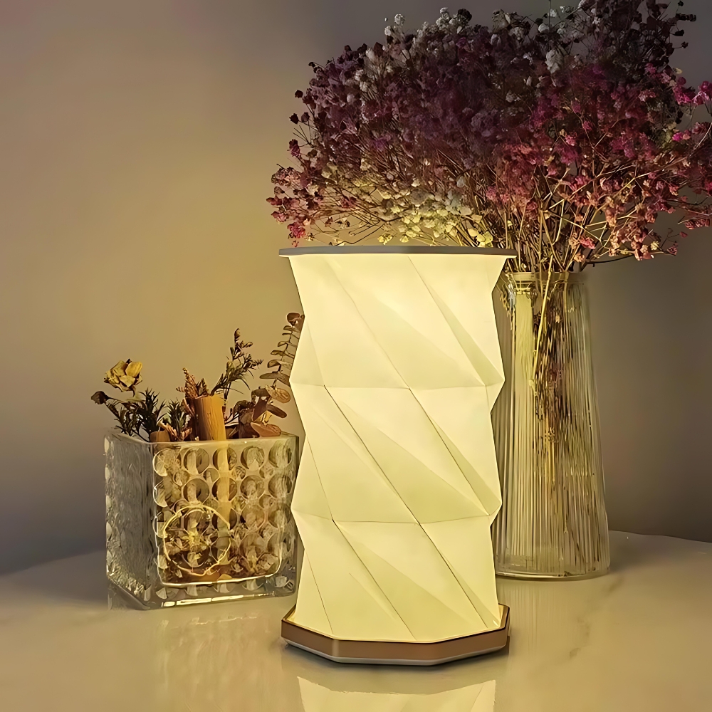 Lampe de chevet LED pliable au design d'origami de couleur beige avec la lumière blanc chaud allumée. Posée sur un support style marbre avec un vase transparent derrière à droite dans lequel se trouvent des fleurs violettes. Derrière la lampe à gauche, un récipient rectangulaire transparent avec des fleurs sèches.