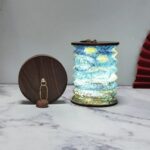 Lampe de chevet sans et pliable de style lanterne japonaise en papier avec un pliage accordéon. Sur chaque extrémité un cercle en bois foncé. Le motif de la lampe ouverte ressemble à une peinture aquarelle d'un paysage bleu et vert. La lampe est allumée. À sa gauche, la même lampe fermée, posée sur la tranche, laissant voir le cercle en bois avec une chaînette dorée. Le tout est posé sur un support style marbre blanc et gris. Le fond est gris.