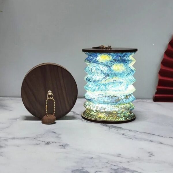 Lampe de chevet sans et pliable de style lanterne japonaise en papier avec un pliage accordéon. Sur chaque extrémité un cercle en bois foncé. Le motif de la lampe ouverte ressemble à une peinture aquarelle d'un paysage bleu et vert. La lampe est allumée. À sa gauche, la même lampe fermée, posée sur la tranche, laissant voir le cercle en bois avec une chaînette dorée. Le tout est posé sur un support style marbre blanc et gris. Le fond est gris.