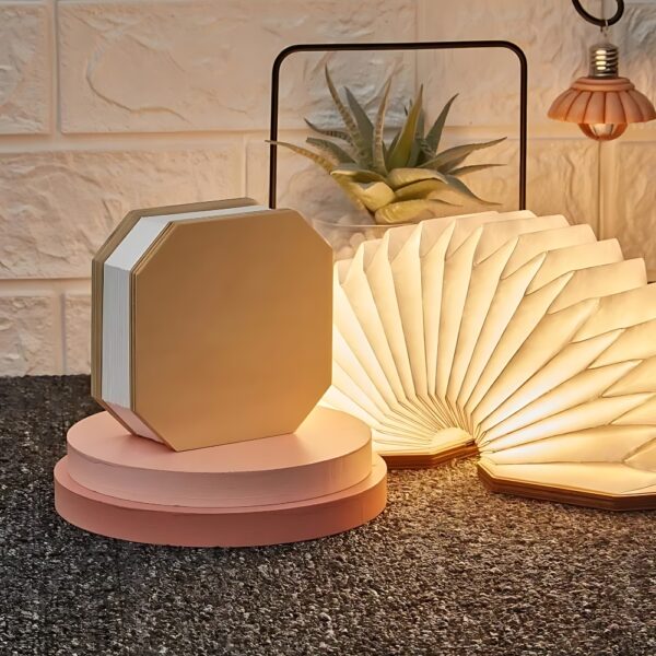 Surface grise sur laquelle sont empilés deux cercles roses et sur lesquels est posée sur sa tranche une lampe sans fil LED pliable effet accordéon fermée. À sa droite, la même lampe ouverte en accordéon et allumée. Derrière elles, une petite plante verte et une suspension rose à droite. Le mur au fond est blanc.