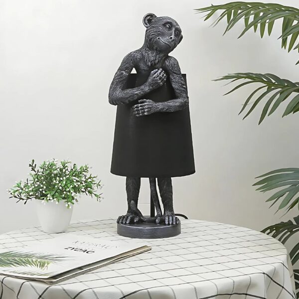 Lampe de chevet jungle singe souriant et câlin, en version noire, sur une table avec une petite plante verte à ses côtés. La table est avec une nappe à motifs dans les tons blanc et vert.