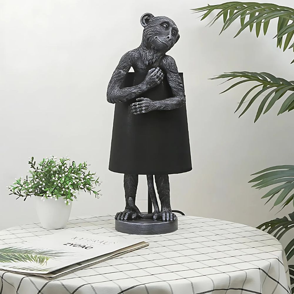 Lampe de chevet jungle singe souriant et câlin, en version noire, sur une table avec une petite plante verte à ses côtés. La table est avec une nappe à motifs dans les tons blanc et vert.