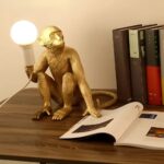 Lampe de chevet singe assis, en version dorée. La lampe est allumée et est posée sur un meuble en bois qui semble être un buffet de salon. La main du singe est posée sur un magazine ouvert et des livres sont rangés derrière cette lampe.