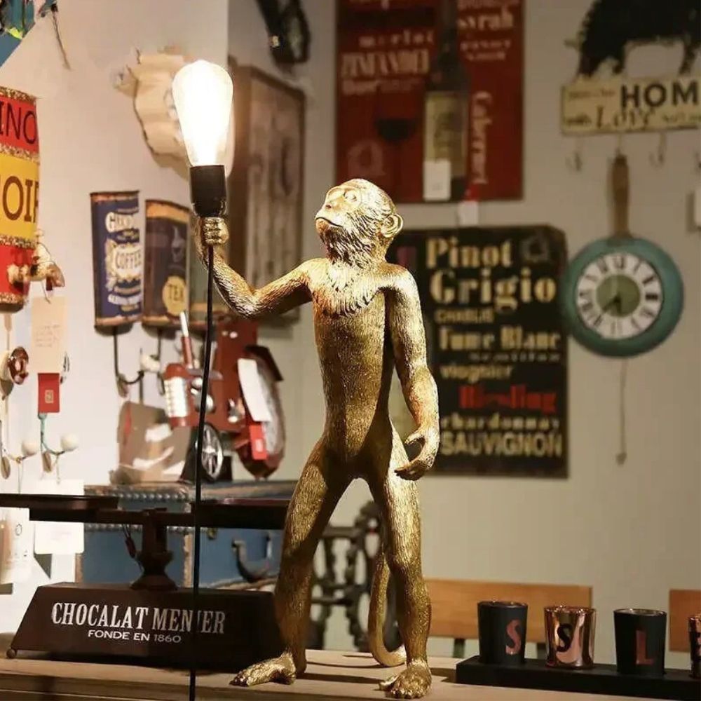 Lampe de chevet singe debout, en version dorée et allumée. Elle est posée sur un bureau et les murs en arrière-plan ont beaucoup de petites affiches en tout genre accrochées dessus.