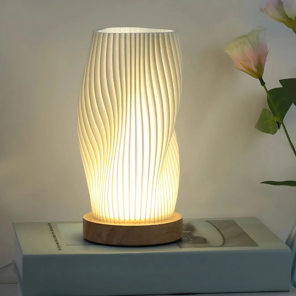 Lampe de chevet élégante avec design ondulé et allumée sur socle en bois. La lampe est posée sur un livre blanc, des fleurs à droite.