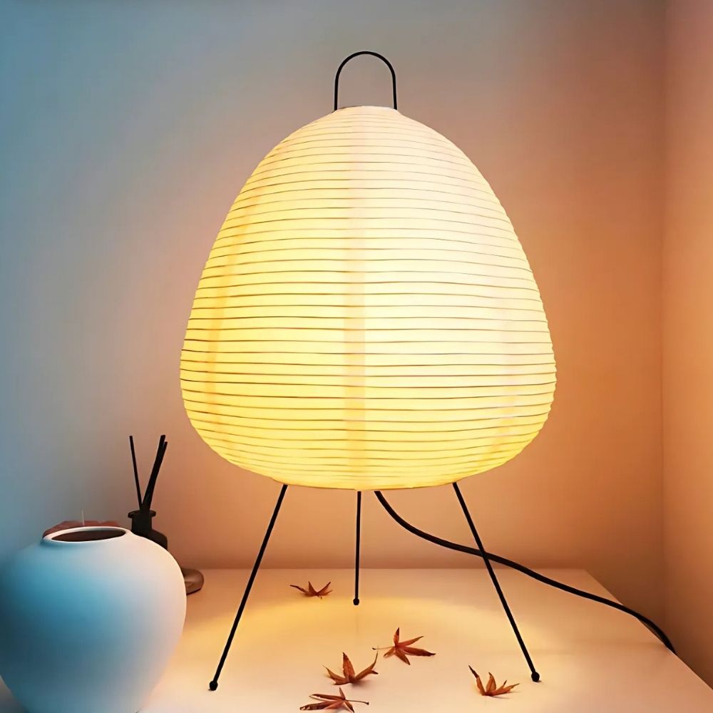 Lampe de chevet japonaise en papier allumée posée sur trois pieds fins en métal posée sur un meuble avec une décoration blanche à gauche.